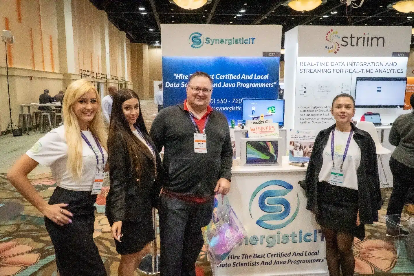 SynergisticIT booth at conference showcasing certified Java and data scientists, with staff and promotional materials.